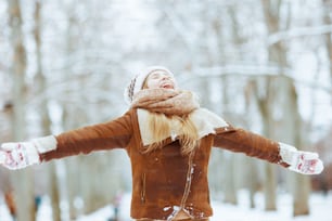 Lächelndes stilvolles Kind in Strickmütze und Schaffellmantel mit Fäustlingen mit offenen Armen freut sich im Winter draußen im Stadtpark.
