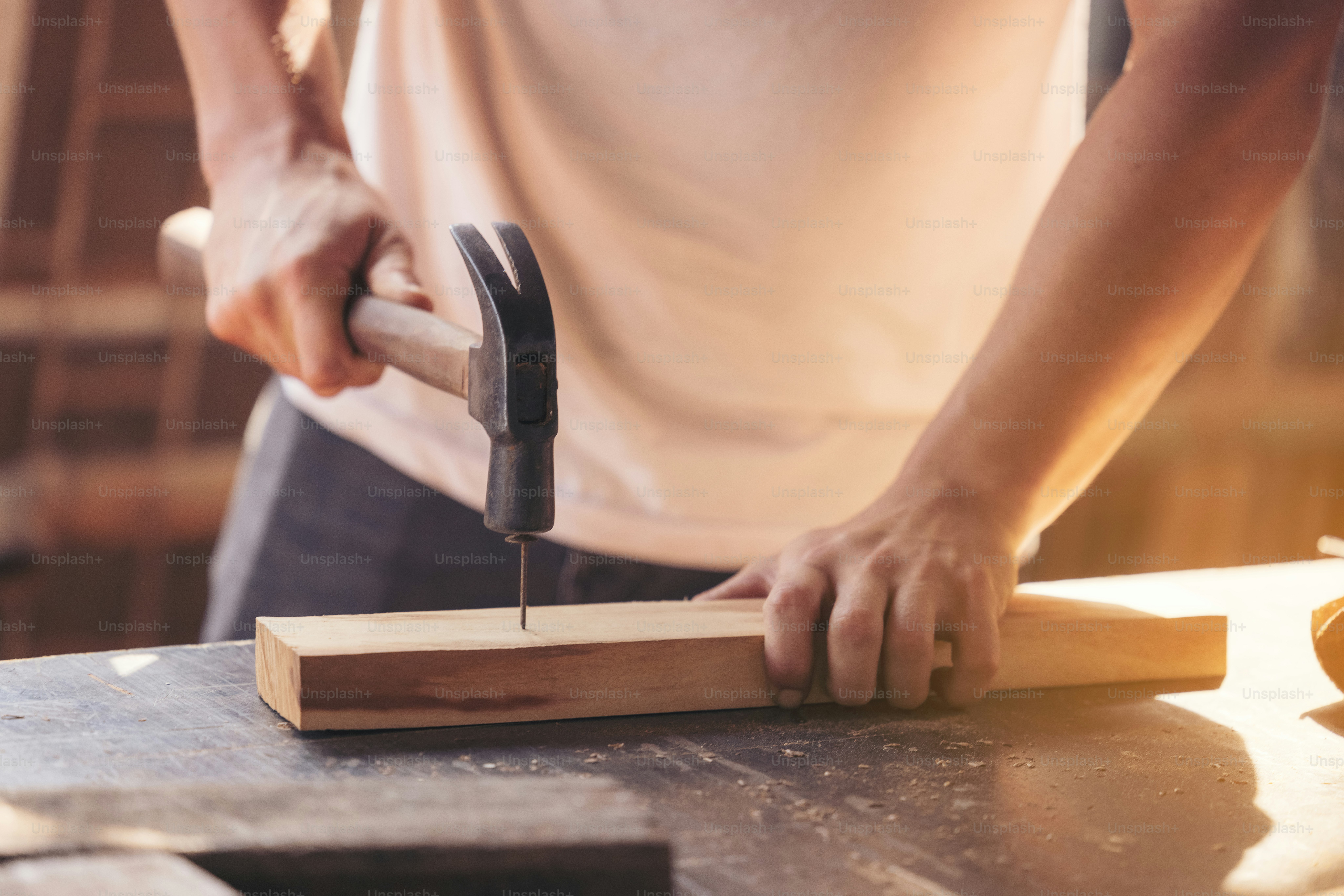 A young male carpenter builder working overall equals a wooden bar with a milling machine in the workshop, in the background wooden