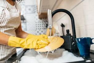 黄色い保護手袋をはめた若い女性が台所の流しで食器を洗っている。家庭および家庭の衛生ルーチン。