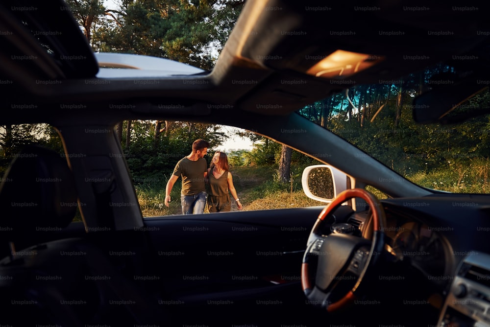 Vista do interior do carro. Volante, espelho lateral. Lindo casal jovem tem um bom tempo na floresta durante o dia.