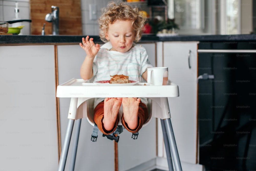 Cute happy Caucasian curly kid boy sitting in high chair eating homemade apple pie and drinking juice. Toddler enjoying food. Healthy eating for kids children. Morning routine breakfast.