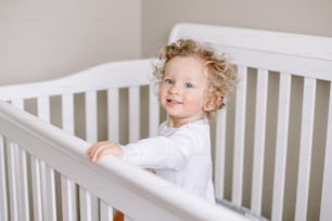 Cute adorable baby boy toddler standing in crib at kids nursery room at home. Funny charming baby boy with curly blond hair and blue eyes playing at his bed. Happy authentic candid home life.