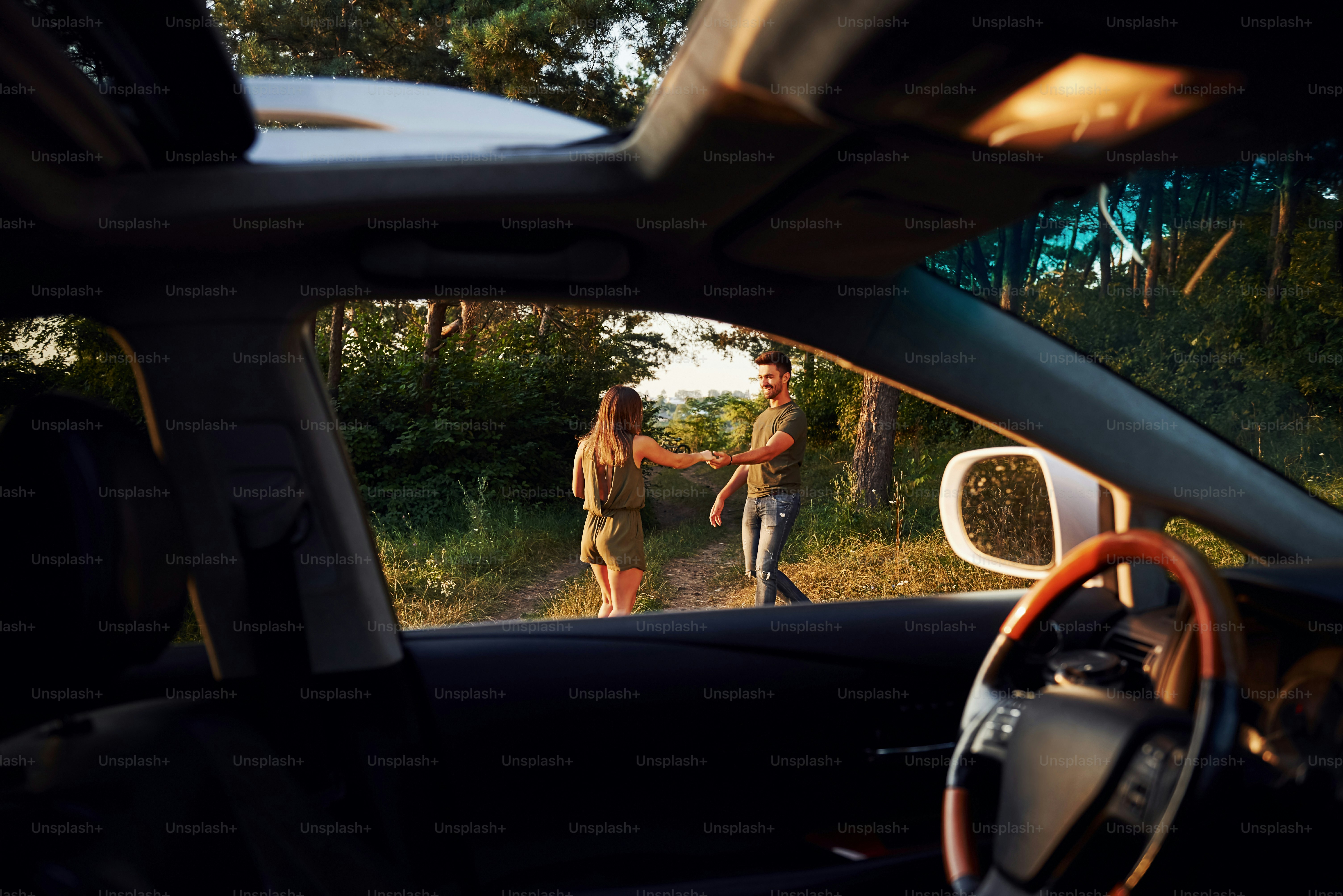 View from the car's interior. Steering wheel, side mirror. Beautiful young couple have a good time in the forest at daytime.