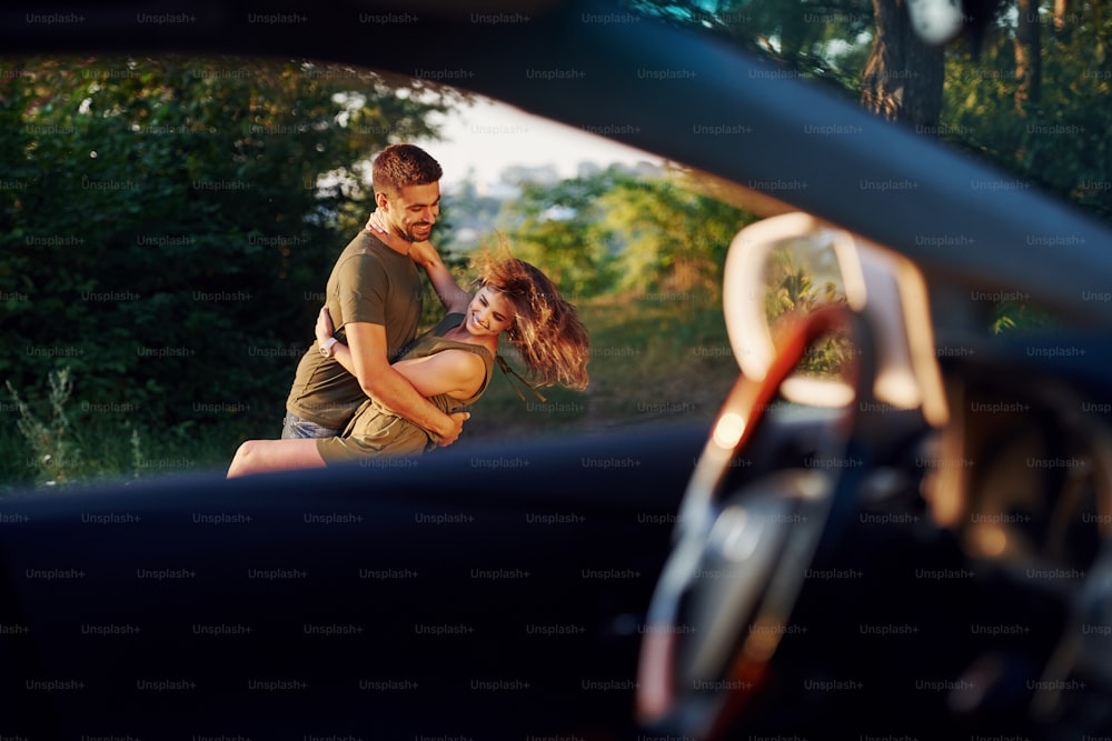 View through the car's window. Beautiful young couple have a good time in the forest at daytime.