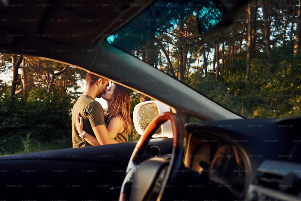車内からの眺め。ステアリングホイール、サイドミラー。美しい若いカップルは、昼間の森で楽しい時間を過ごしています。