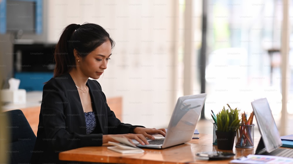 Junge asiatische Geschäftsfrau, die an ihrem Projekt mit einem Laptop arbeitet, während sie an einem Schreibtisch sitzt.