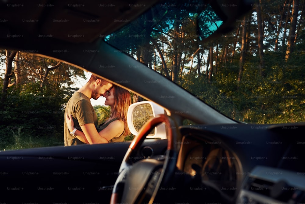 View from the car's interior. Steering wheel, side mirror. Beautiful young couple have a good time in the forest at daytime.