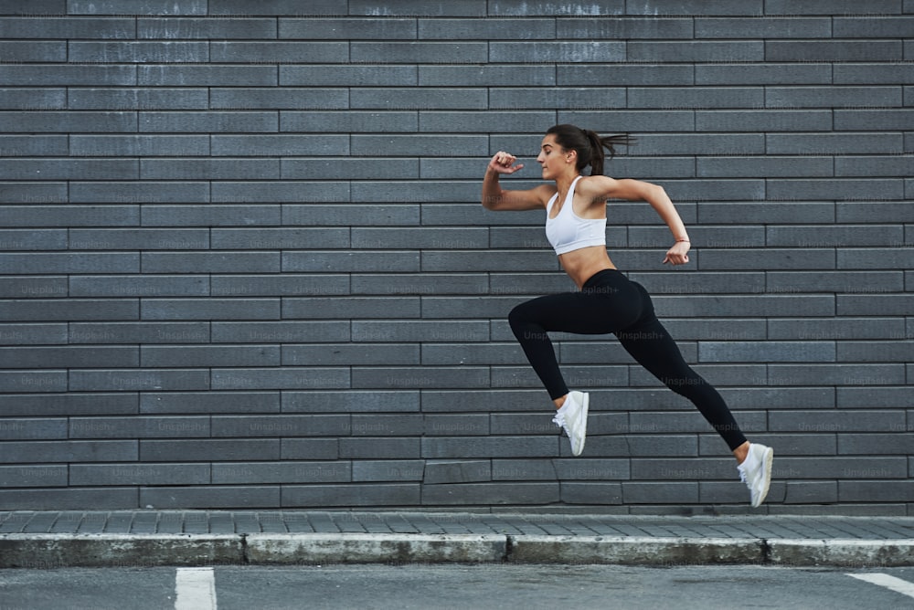 Running fast. Young sportive brunette with slim body shape against brick wall in the city at daytime.