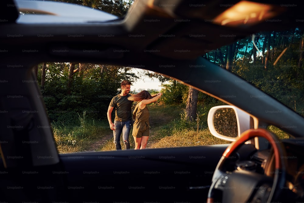 車内からの眺め。ステアリングホイール、サイドミラー。美しい若いカップルは、昼間の森で楽しい時間を過ごしています。