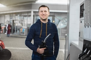 Un jeune homme caucasien vêtu d’une tenue décontractée, est prêt à faire le plein de sa voiture de luxe, tenant la buse du pistolet de remplissage à la station-service, regardant la caméra avec le sourire