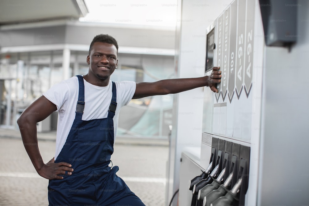 Handsome young smiling black gas station worker on white t-shirt and blue overalls, posing to camera, while leaning on the panel with filling guns nozzles at modern petrol station.
