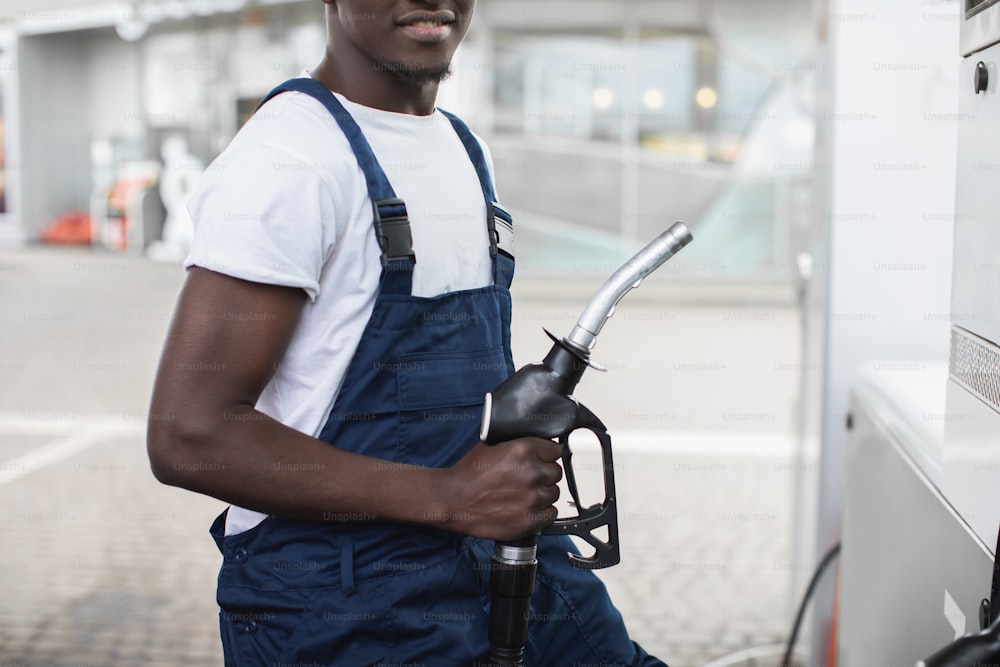 Close up cropped image of black skinned man in workwear, worker of petrol station, standing outdoors at the petrol station and holding nozzle of fuel gun in hands.