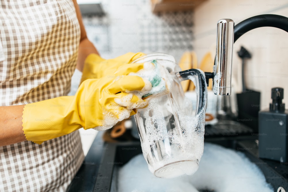 Giovane donna adulta con guanti protettivi gialli che lava i piatti sul lavello della cucina. Routine di igiene domestica e domestica.