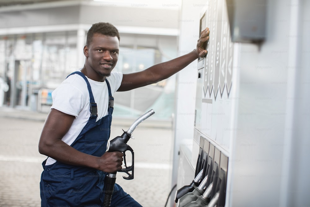 Junger, lächelnder, angenehmer afrikanischer Mann, Tankstellenarbeiter in Arbeitskleidung, posiert vor der Kamera, während er an der Tankstelle steht, bereit, das Auto mit der Tankpistole zu betanken.