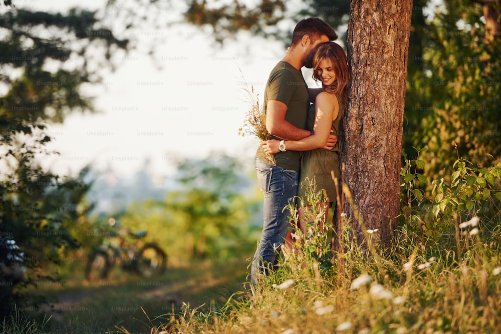 Leaning on the tree. Beautiful young couple have a good time in the forest at daytime.