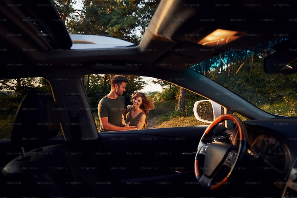 Vista dall'interno dell'auto. Volante, specchietto laterale. Bella giovane coppia si diverte nella foresta durante il giorno.