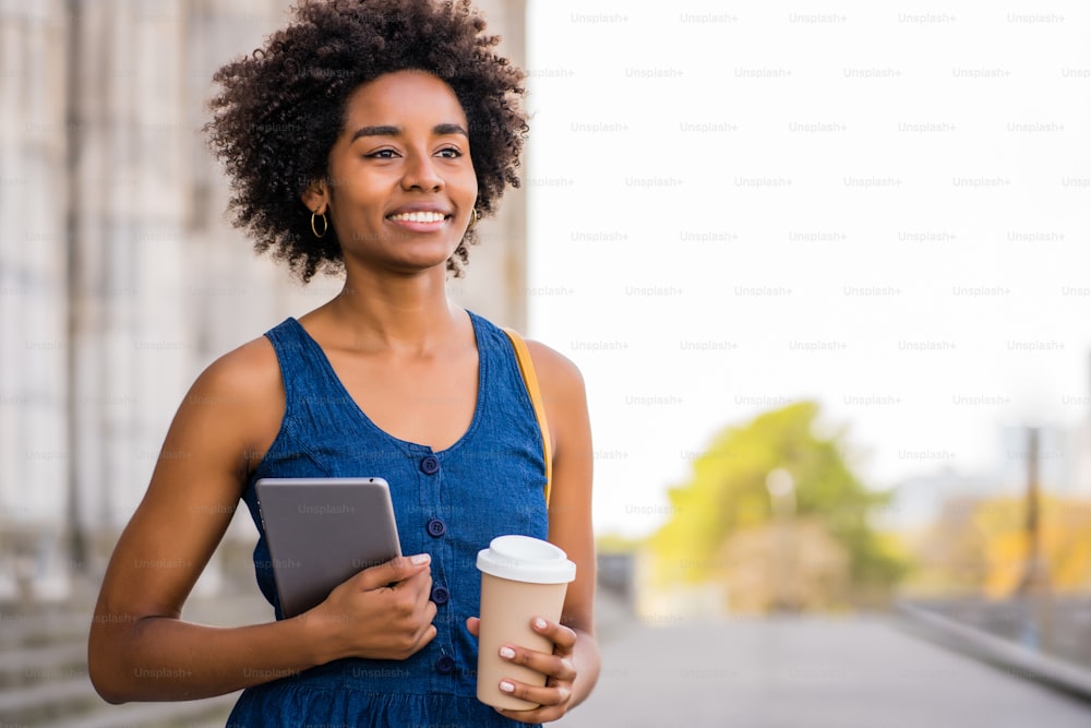 Retrato de una mujer de negocios afro sosteniendo una tableta digital y una taza de café mientras está de pie al aire libre en la calle. Concepto empresarial y urbano.