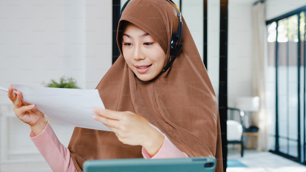 La signora musulmana asiatica indossa le cuffie utilizzando il tablet digitale parla con i colleghi del rapporto di vendita in videochiamata in conferenza mentre lavora da casa in cucina. Distanziamento sociale, quarantena per il corona virus.