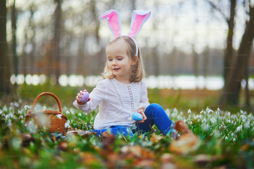 Muchacha con orejas de conejo jugando a la caza de huevos en Pascua. Niño pequeño sentado en la hierba con muchas flores de campanilla de invierno y recogiendo huevos de colores en una canasta. Niño pequeño celebrando la Pascua al aire libre