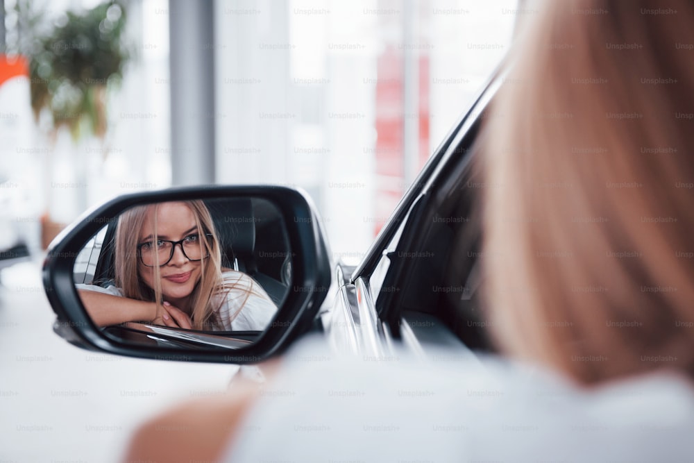Foto enfocada. Mujer con gafas mirando por el espejo lateral de un vehículo moderno en el salón.