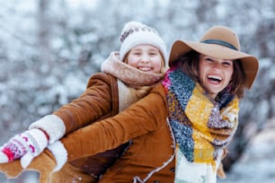 Felice madre elegante e bambino in cappelli e cappotti di pelle di pecora con guanti che giocano fuori nel parco cittadino in inverno.