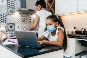 Niña pequeña que tiene una sesión de aprendizaje en línea durante la crisis de la pandemia de Covid-19, el bloqueo o la cuarentena. Madre ocupada trabajando en la cocina y preparando el almuerzo en el fondo. Prevención de enfermedades y concepto de nueva normalidad.