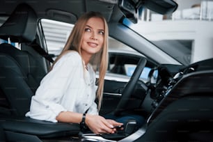 Holds smartphone and smiling. Beautiful blonde girl sitting in the new car with modern black interior.