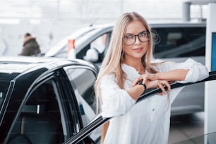 Linda chica con gafas se para cerca del coche en el salón de automóviles. Probablemente su próxima compra.