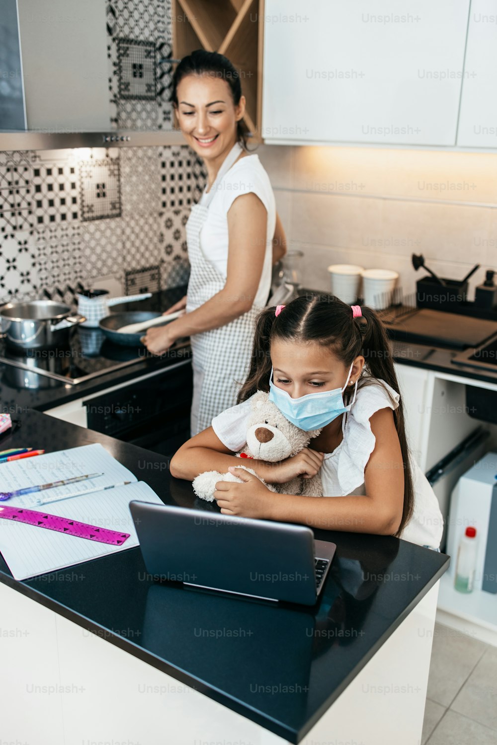 Menina tendo sessão de e-learning durante lockdown ou quarentena da crise da pandemia de Covid-19. Mãe ocupada trabalhando na cozinha e preparando o almoço ao fundo. Prevenção de doenças e novo conceito normal.