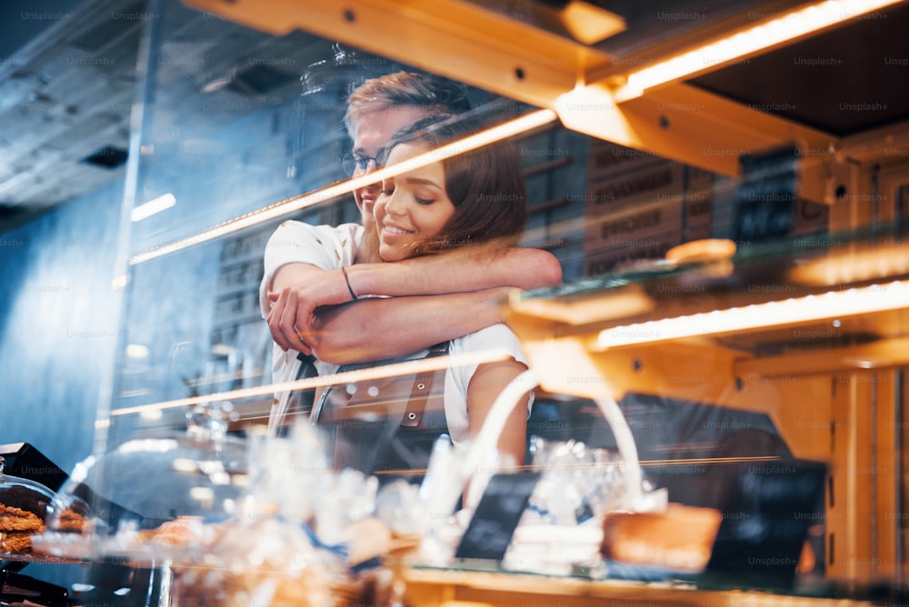 Embracing each other. Behind the glass. Light stripes. Two young cafe workers indoors. Conception of business and service.