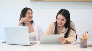 Two young woman designer in casual wear laughing while working with laptop and digital tablet at modern office.