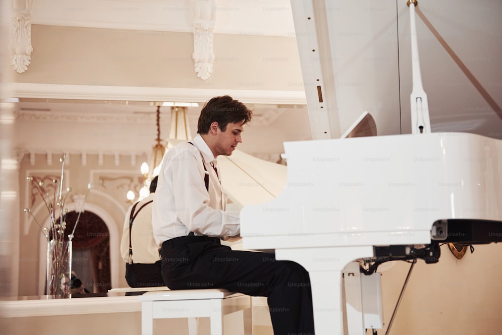 Jovem músico profissional em roupas oficiais toca no piano branco.