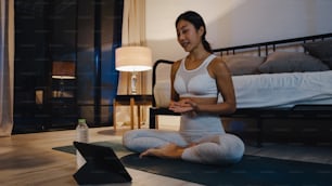 Giovane donna asiatica in esercizi di abbigliamento sportivo che si allena e utilizza il tablet per guardare il video tutorial di yoga a casa sera. Formazione a distanza con personal trainer, distanziamento sociale, concetto di formazione online.