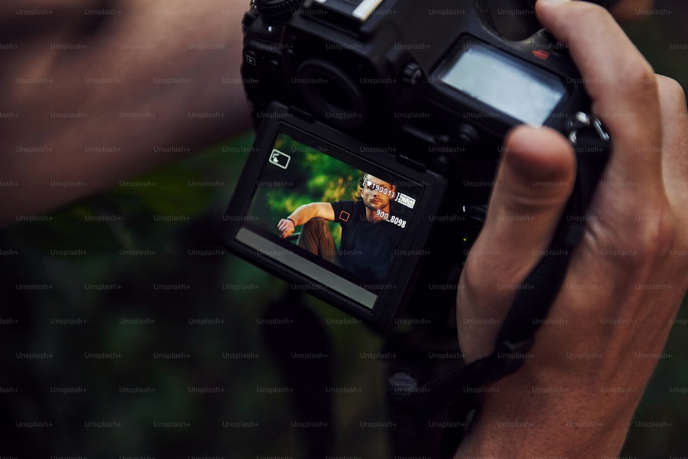 Les mains du photographe tiennent l’appareil photo. Dans les coulisses d’une séance photo en forêt. Homme en chemise noire.
