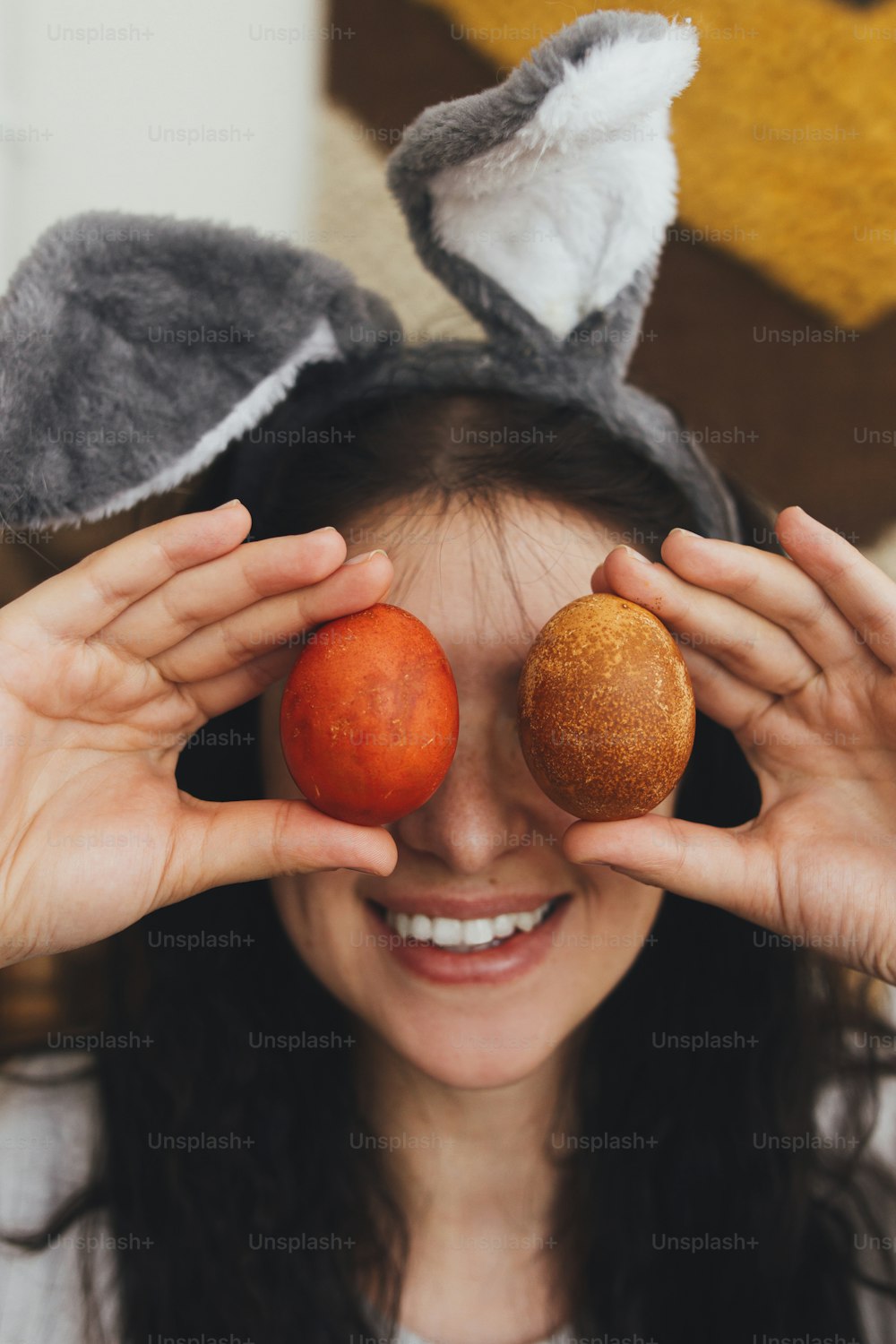 Happy Easter! Happy young woman in bunny ears and linen dress holding easter eggs at eyes and smiling on background of rustic room. Natural dyed easter eggs in hands, funny moments