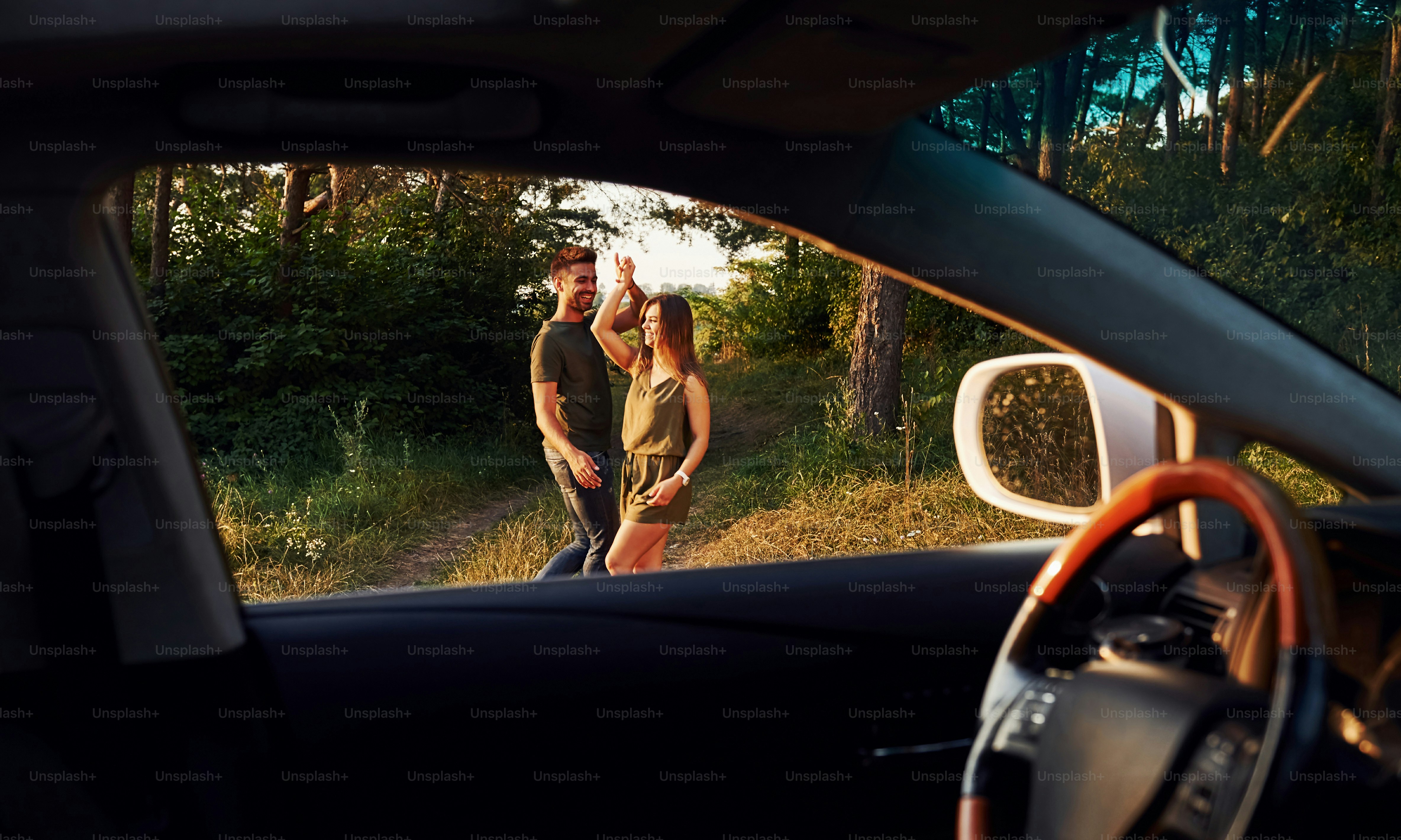 View from the car's interior. Steering wheel, side mirror. Beautiful young couple have a good time in the forest at daytime.