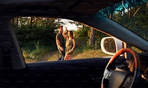 View from the car's interior. Steering wheel, side mirror. Beautiful young couple have a good time in the forest at daytime.