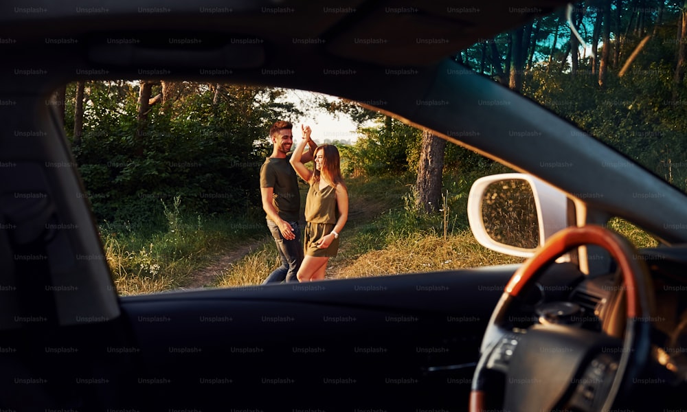 View from the car's interior. Steering wheel, side mirror. Beautiful young couple have a good time in the forest at daytime.