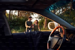 Blick aus dem Innenraum des Autos. Lenkrad, Seitenspiegel. Schönes junges Paar hat tagsüber eine gute Zeit im Wald.