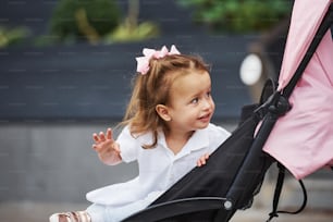 Portrait of cheerful little girl sitting in the pink pram while have walk in the city.