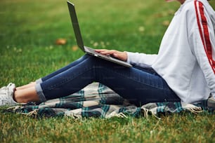 Vista de partículas. Chica positiva con ropa informal con su computadora portátil se sienta en la hierba verde.