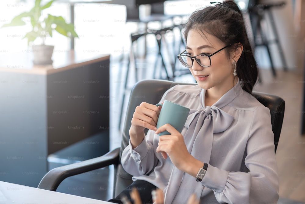 La giovane donna d'affari asiatica ha il piacere di guardare il lavoro sul proprio laptop e il caffè preferito in ufficio.