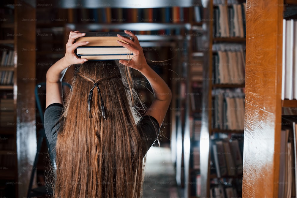 쾌활한 소녀는 그녀의 머리에 책을 넣는 동안 재미를 느낍니다. 여학생이 도서관에 있습니다. 교육의 개념.