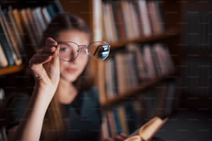 Fokussiertes Foto. Hält die Brille in der Hand. Studentin ist in der Bibliothek, die voller Bücher ist. Konzeption von Bildung.