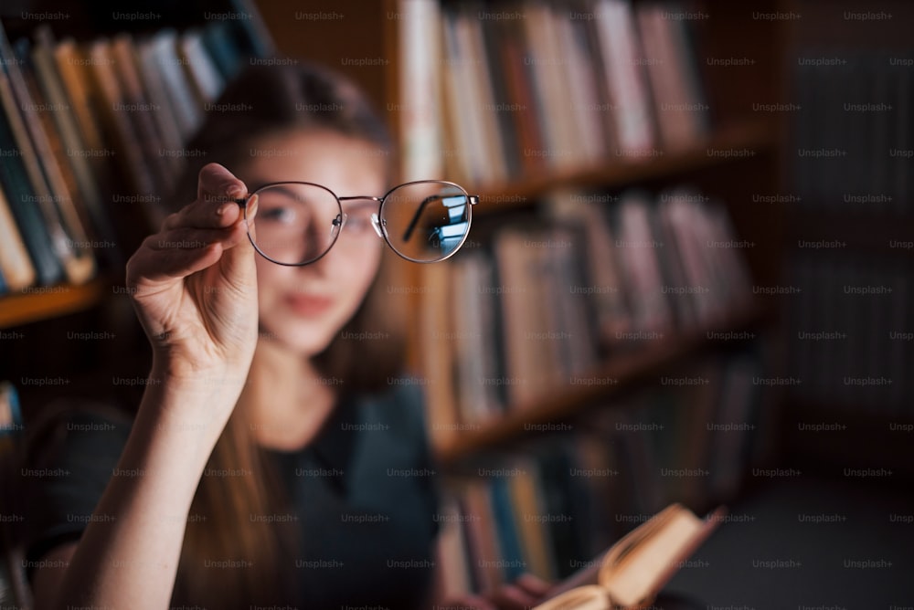 Focused photo. Holds glasses in hand. Female student is in library that full of books. Conception of education.
