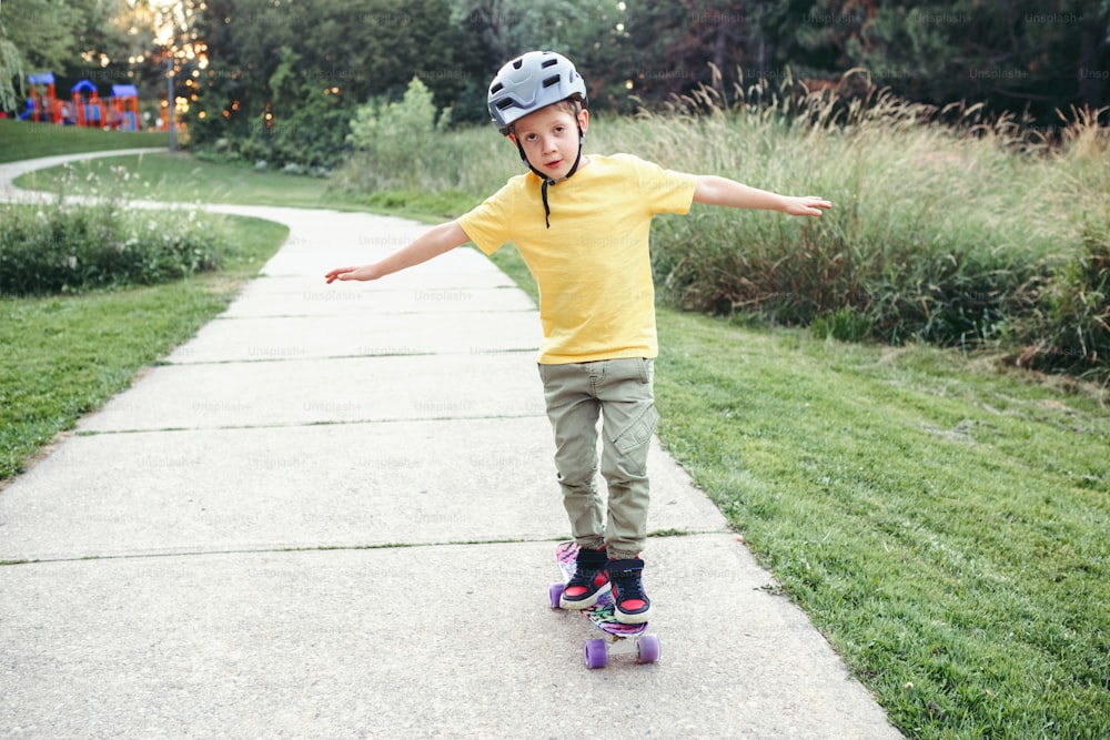 Happy Caucasian boy in grey helmet riding skateboard on road in park on summer day. Seasonal outdoor children activity sport. Healthy childhood lifestyle. Boy learning to ride skateboard.