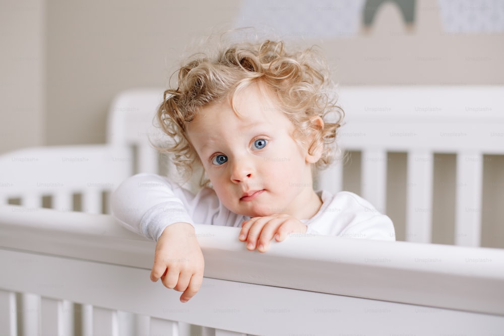 집에 있는 어린이 보육실의 유아용 침대에 서 있는 귀여운 사랑스러운 남자 아이. 곱슬곱슬한 금발 머리와 카메라를 바라보는 파란 눈을 가진 호기심 많은 매력적인 아기. 행복한 정통 솔직한 가정 생활.