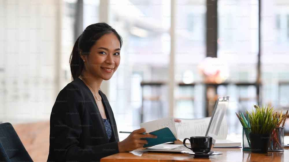 Confident businesswoman holding notebook and smiling to camera while sitting at office desk.