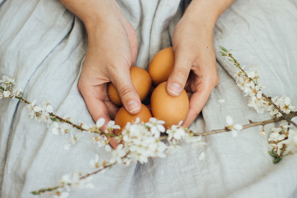 Happy Easter! Easter eggs and spring flowers in hands of woman in rustic linen dress. Woman holding natural easter eggs and spring blooms. Aesthetic eco holiday, stylish image