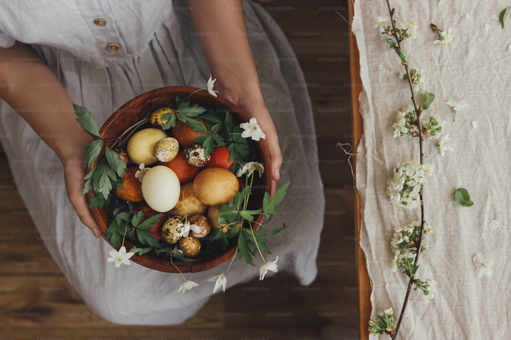 Woman in linen dress holding easter eggs and spring flowers in wooden bowl at table, top view. Stylish easter and quail eggs in natural dye and spring blooms. Aesthetic holiday. Happy Easter!